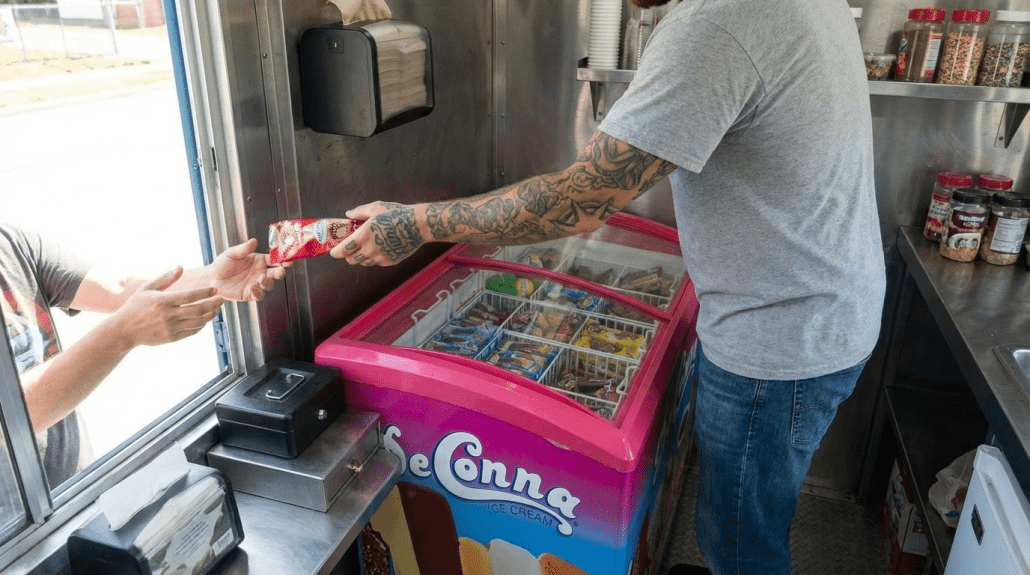Ice cream truck owner handing a DeConna ice cream novelty to a customer from a pink sliding-glass freezer stocked with treats.