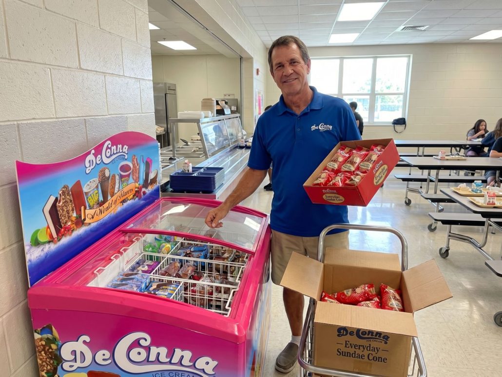 School Cafeteria Ice Cream Freezer Restocking - DeConna Ice Cream Smiling man in a blue polo stocks a pink DeConna ice cream chest in a school cafeteria.