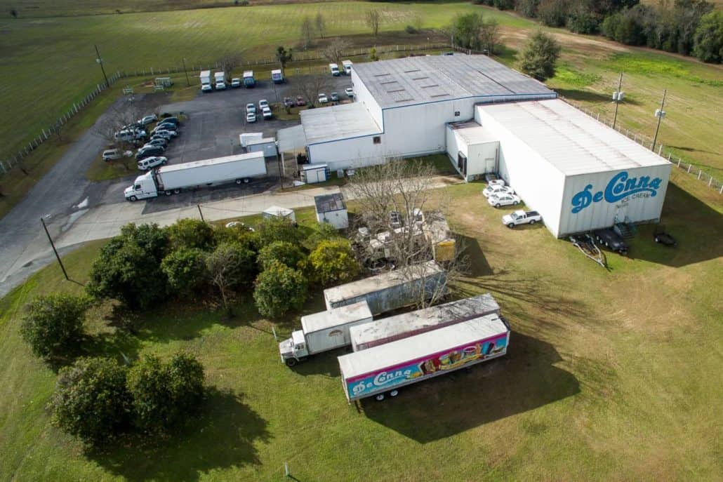 Aerial view of an ice cream factory complex with white buildings, loading docks, and multiple trucks parked in a lot surrounded by green fields.