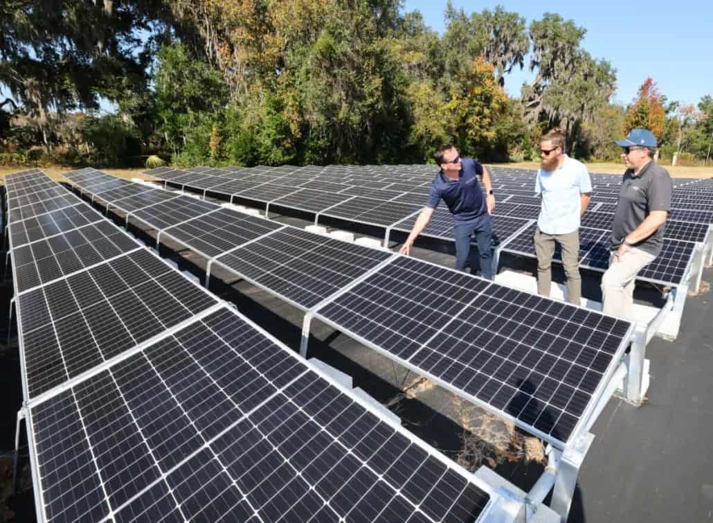 Three men inspect a large solar panel array outdoors, pointing at panels amid a sunny, tree-filled background.