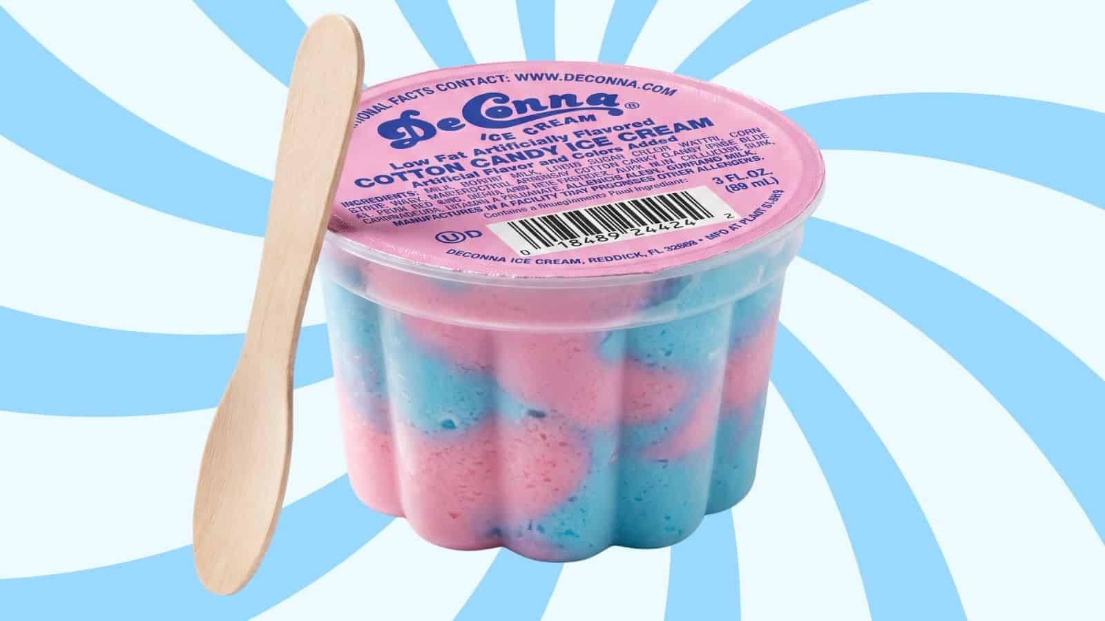 Container of pink-and-blue cotton-candy ice cream with a wooden spoon on the side, pink lid labeled DeConna, set against a blue-and-white swirl background.
