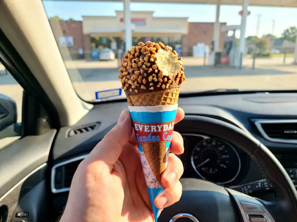 Hand holding a DeConna Everyday Sundae Cone with its wrapper pushed down, in a car outside a convenience store