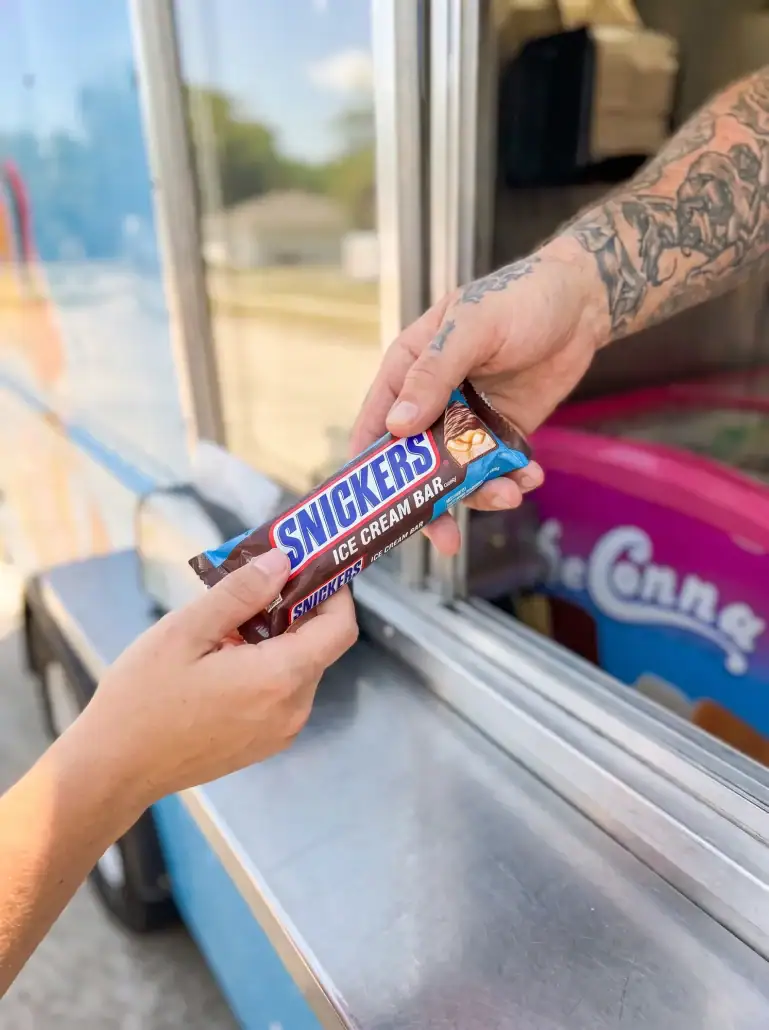Snickers Ice Cream Bar at the Truck Window (Portrait) - DeConna Ice Cream Vertical close-up of a sealed Snickers ice cream bar handed through an ice cream truck window, DeConna freezer behind